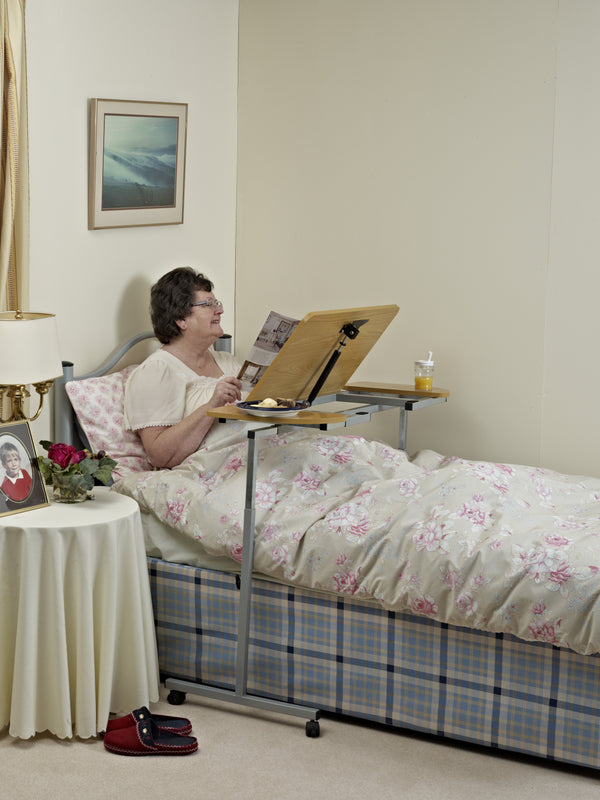 Image shows an individual in bed, reading from a titled over bed desk. The room is mainly cream, with a landscape framed image above the bed, and the bedding is floral cream and pink. 