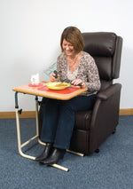 This photo shows an individual sat in a brown leather arm chair, with the wooden desk set up over their lap with a mug and bowl of food on it from which they are eating. The carpet is grey, and the walls are white with wooden skirting boards. 