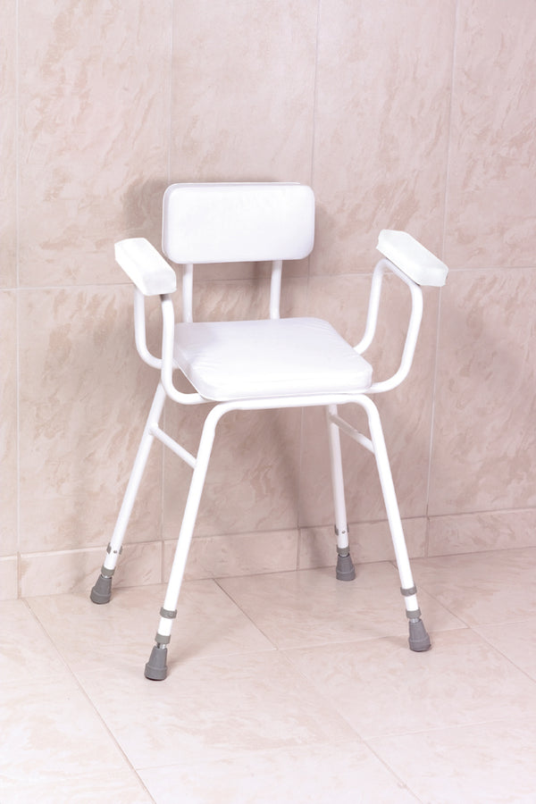 This image shows the white chair against a cream tiled wall and floor. The chair has all white frame, two cushioned arm rests, cushioned seat and back support, and four legs.