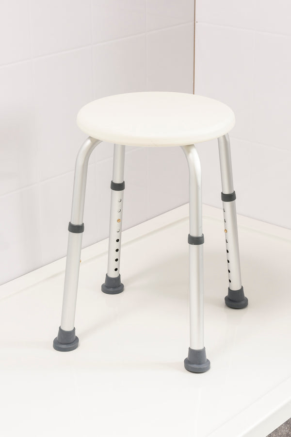 This image shows the shower stool, with white round seat and silver legs, with grey grips at the bottom, in a white shower.