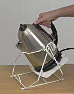 Demonstrating how to use the product, with a silver kettle in white kettle tipper. The wall is pale grey and the surface its on is wooden. 