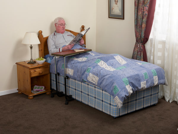 An individual in bed reading, using the Brown Overbed/ chair table. The single bed has blue covers, and is in a cream room next to a wooden bedside table with a cream lamp. 