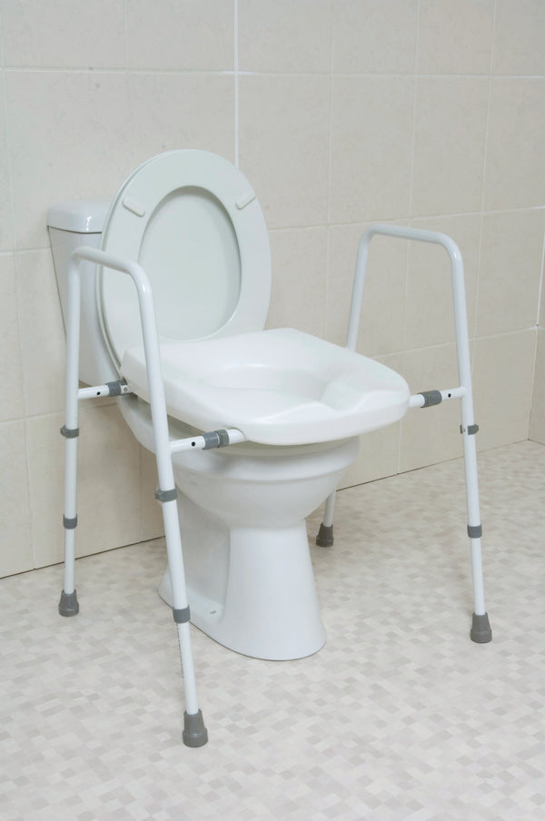 The image shows the white toilet frame and toilet seat set over a white toilet, in a cream bathroom. The grey sections on the frame show the width adjustment and the grips on the feet. 