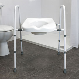 This image shows the assembled white toilet frame and seat. The product is shown in the middle of a bathroom with grey tiled floors, white tiled walls and has a white toilet and white shower in the background. 
