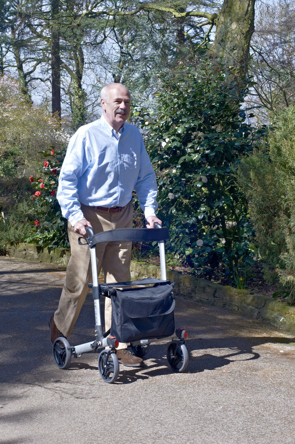 Image shows a person pushing the silver rollator outside on a path. There are lots of trees and bushes beside the path. 