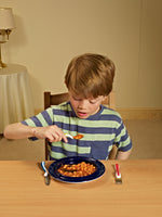 Image shows a child eating baked beans from a plate using the Kura Care children's cutlery set, at a wooden table, in a beige room. 