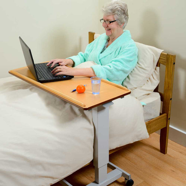 Image shows an individual in bed, with a laptop and drink on a brown desk with grey metal frame across the bed. The walls and bedding are cream and the floor and furniture are wooden. The individual is wearing a mint green dressing gown. 