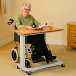 This image shows an individual in an olive green long sleeved top sat in a black wheelchair. They are using the table to read, write and access water and food comfortably. They are sat in a cream room with wooden furniture. 