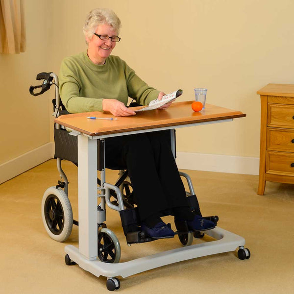 This image shows an individual in an olive green long sleeved top sat in a black wheelchair. They are using the table to read, write and access water and food comfortably. They are sat in a cream room with wooden furniture. 