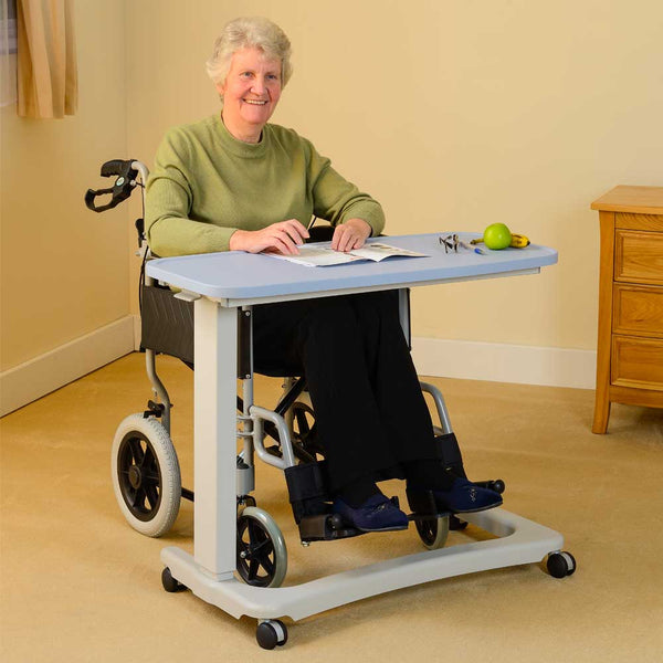 This image shows an individual in a long sleeved olive green top, sat in a black wheelchair, with the blue desk pulled over it so they can read and write easily. They are sat in a room with cream walls and carpet and a wooden chest of drawers. 