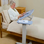 This image shows an individual sat up in bed with the tilted pale blue over-bed desk set at a comfortable angle for them to read and write. The room and bedding are cream with wooden furniture. 