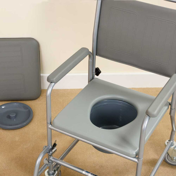 This image shows the grey commode from a higher angle to show the depth of the commode pan. The commode is in a beige and cream room and the pan's lid, and the removable cushion to cover the commode, are set on the floor against the wall. 