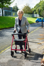 This image shows an individual walking outside, using the red rollator on a concrete path, with a green bank and buildings behind them. 