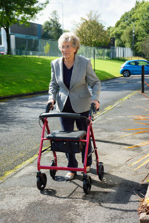 This image shows an individual walking outside, using the red rollator on a concrete path, with a green bank and buildings behind them. 
