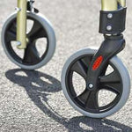 Image shows two of the champagne Rollators wheels, which are black with grey rims, on concrete ground. 